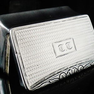 May include: A silver hinged box with a textured surface and a floral design on the bottom edge. The box is engraved with the initials "T.T."