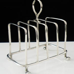 May include: A silver-toned metal toast rack featuring four vertical dividers and a central loop handle. The structure is supported by four small feet shaped like animal paws, providing a stable base for holding slices of toast upright.