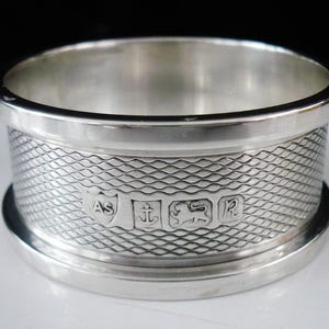 May include: A silver napkin ring with a textured, wave-like pattern around the center. The ring has a smooth, polished rim and base. The center of the ring features several engraved symbols.