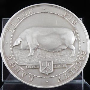 May include: A silver coin featuring a detailed relief of a pig standing on grass. The coin's outer ring reads "LARGE BLACK PIG SOCIETY" and includes the text "Founded 1899" and the letters "LBP".