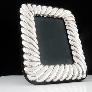 May include: Silver-toned picture frame with a rope-like border. The frame has a rectangular shape with rounded corners and a dark interior. The frame is displayed on a white surface against a black background.