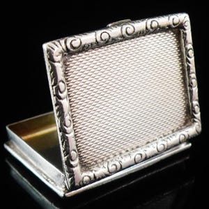 May include: A small, open silver box with a rectangular shape. The lid features a textured, patterned surface and an ornate, scrolled border. The interior of the box has a gold-colored finish. The box is set against a black background.