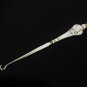 May include: A silver-toned button hook with an ornate handle and a curved hook end. The handle features decorative filigree and is topped with a small, amber-colored gem. The hook is designed for fastening buttons on clothing.