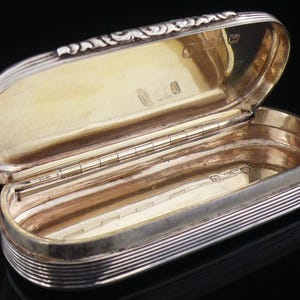 May include: An oval silver box with a hinged lid, featuring a ribbed exterior and a gold-toned interior. The lid edge displays decorative embossed scrollwork, and the interior surfaces are marked with small, clear hallmarks on both the base and lid.