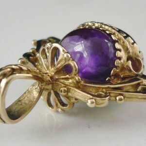 May include: A gold-toned pendant featuring a large, round, deep purple gemstone. The pendant has an ornate, filigree design with a loop for a chain. The gemstone is set in a decorative bezel.