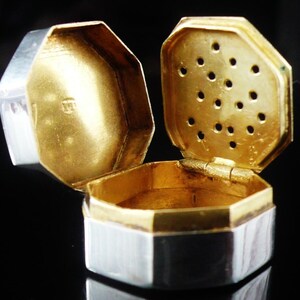 May include: An open, octagonal antique box with a hinged lid. The box is gold-coloured on the inside and has a silver and white exterior. The lid has small holes. The box is likely made of metal and is displayed against a black background.