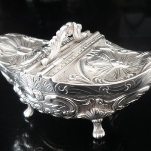 May include: A silver hinged box with ornate floral designs and three feet. The box has a decorative handle on the top.