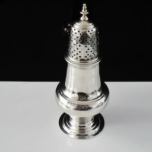 May include: A silver sugar caster with a pierced top and a decorative finial. The caster has a bulbous body and a round base. The background is black and white. The sugar caster is likely made of sterling silver.