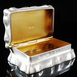 May include: A rectangular silver box with a hinged lid and a gold-toned interior. The exterior features a wavy, undulating surface texture. The inner lid and rim display small stamped hallmarks and markings visible against the metallic gold finish.
