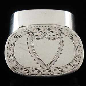 May include: A silver oval box with a hinged lid. The lid is decorated with an engraved shield design within an oval frame, surrounded by detailed floral and leaf patterns. The box has a polished, reflective surface.