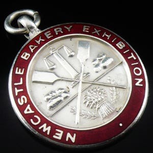 May include: A silver-toned round medal with a red border, featuring the text "Newcastle Bakery Exhibition." The center displays crossed baking tools and wheat. A loop is at the top for hanging.