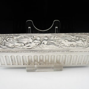 May include: A silver rectangular box with an ornate design featuring a scene of people and animals. The box has a ribbed bottom.
