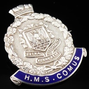 May include: A silver-toned metal badge featuring a central shield with an eagle emblem, surrounded by a wreath and a crown at the top. A blue banner at the bottom displays the text H.M.S. COMUS, while smaller banners read CHINA, COPENHAGEN, and HORNS REEF.