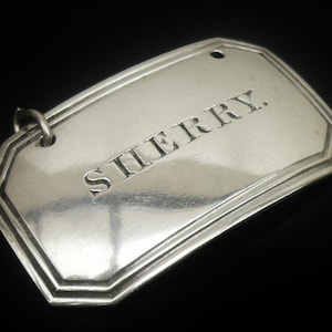 May include: A silver tag with the word "SHERRY" engraved on it. The tag has a rectangular shape with a decorative border and a small hole at the top for hanging. The background is black.