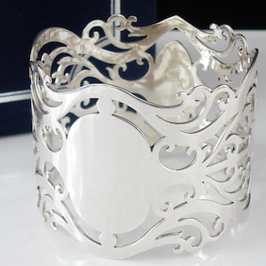May include: A silver napkin ring with an ornate, openwork design. The ring features a repeating pattern of swirling lines and curves, with a smooth, oval space for engraving. The metal has a bright, reflective finish.