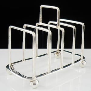 May include: A silver-toned metal toast rack featuring four rectangular slots for holding slices of bread. The structure is supported by four small spherical feet at the base and includes a central handle loop for carrying the rack easily.