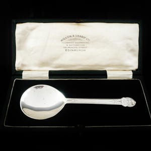 May include: A silver spoon with a rounded bowl and a detailed handle, presented in a black box with a cream-colored satin lining. The box is open, revealing the spoon's polished surface. The spoon is likely a collectible or antique item.