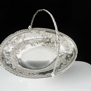 May include: A silver-colored, oval-shaped basket with a twisted handle. The basket features ornate floral and grape vine detailing around the rim and on the interior. The handle is also silver and twisted. The basket is on a white surface with a black background.