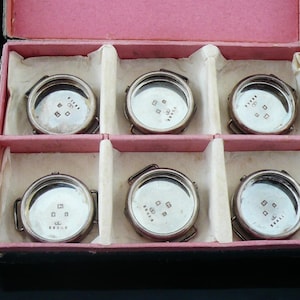 May include: Six small, circular, silver-coloured watch components, each with a clear face and small markings. They are presented in a pink and white divided box. The box is open, displaying the contents.
