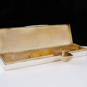 May include: A rectangular silver-toned box with a hinged lid, open to reveal a gold-toned interior with compartments. A gold-toned rectangular piece with a handle and a small pair of scissors are on the right side. The box is on a white surface.