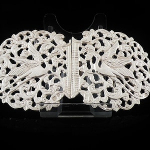 May include: Silver-colored, ornate buckle with a symmetrical design. Two stylized birds are featured within a detailed, openwork pattern of swirling leaves and vines. The buckle is displayed against a black background.