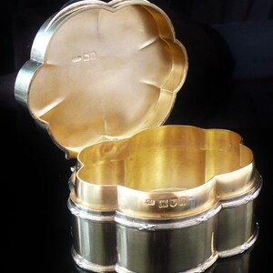 Könnte beinhalten: Eine goldfarbene, kleeblattförmige Schatulle mit Scharnierdeckel. Die Schatulle hat ein silberfarbenes Band um den Boden und den Deckel. Das Innere der Schatulle ist goldfarben. Die Schatulle ist geöffnet und gibt den Blick ins Innere frei.