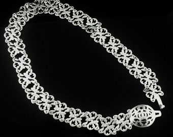 May include: A silver-colored filigree belt with an ornate floral design. The belt is made of interconnected, repeating floral patterns. The clasp is a circular design with a decorative floral motif. The belt is displayed against a black background.