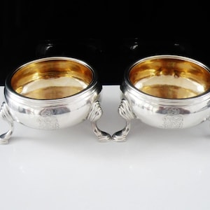 May include: A pair of round silver bowls with gold-toned interiors, each featuring three curved feet and an engraved crest on the exterior side. The bowls have a polished metallic finish and a wide, open top design, standing on a smooth surface.