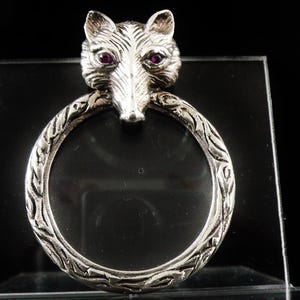 May include: A silver-coloured magnifying glass featuring a wolf head. The wolf has red gemstone eyes and sits above a circular frame with an ornate, textured pattern. The magnifying glass is displayed on a clear, rectangular stand.