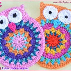 Op de afbeelding: Twee gebreide uil-onderzetters, een roze en een oranje, met kleurrijke mandala-ontwerpen in het midden. De uilen hebben witte ogen met zwarte pupillen en roze snavels.