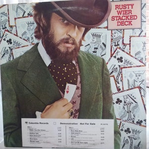 May include: A man in a green suit and a black hat holds an ace of hearts playing card in his pocket. The background is a collage of playing cards. The album cover text reads "Rusty Wier Stacked Deck" and "Columbia Records Demonstration Not For Sale PC 34778".