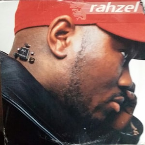 May include: A close-up of a man's face, wearing a red baseball cap with the word "rahzel" printed on it. He is wearing a black leather jacket and has a small electronic device attached to his neck.
