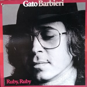 May include: Album cover featuring Gato Barbieri in a black hat and glasses. The album title "Ruby, Ruby" is displayed in pink text at the bottom. The artist's name, "Gato Barbieri," is at the top.