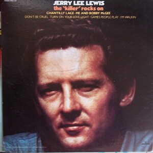 May include: A vintage record album cover featuring Jerry Lee Lewis. The cover is a close-up of Lewis's face, with a dark background. The album title, "the 'killer' rocks on", is printed in white letters at the top of the cover. The album's track list is printed below the title.