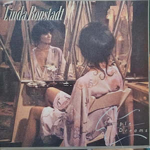 May include: A vintage vinyl record album cover featuring Linda Ronstadt. The cover art shows a woman in a pink robe sitting in a chair, with a reflection of her in a mirror behind her. The album title "Simple Dreams" is written in cursive on the bottom right corner.