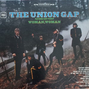 May include: Album cover for "The Union Gap" featuring Gary Puckett. The album cover has a blue and white title with the words "Woman, Woman" in green. The cover features a group of men in black uniforms standing in a forest setting.