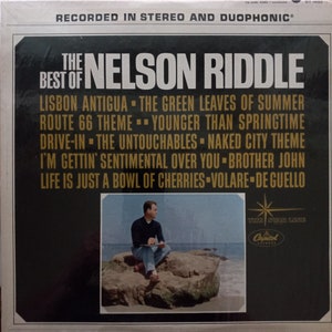 May include: A black and white vinyl record cover with a gold border. The cover features a photo of a man sitting on a rock by the ocean. The text on the cover reads "The Best of Nelson Riddle" and lists the songs included on the record. The Capitol Records logo is in the bottom right corner.