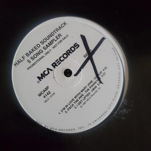 May include: A white vinyl record with the text "HALF BAKED SOUNDTRACK 5 SONG SAMPLER" and "MCA RECORDS". The record has a large black "X" drawn on it. The record includes song titles and artist names.