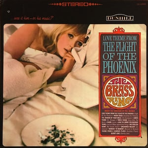 May include: A vintage vinyl record album cover featuring a woman lying in bed with a plate of cigarettes on the nightstand. The album cover is for "The Brass Ring" and features the title "Love Theme From The Flight Of The Phoenix".