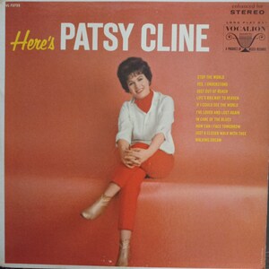 May include: A vinyl record cover titled Here's Patsy Cline with a red background. The cover features a photograph of a person with dark hair wearing a white shirt, red turtleneck, and red pants, paired with tan boots. A tracklist is printed in yellow.