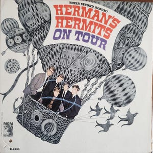 May include: Album cover for "Herman's Hermits On Tour." The cover art features a whimsical illustration of the band in a hot air balloon, with the band's name in red and purple text. The background is white with gray and black accents.