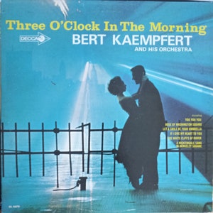 May include: A vintage vinyl record cover featuring a silhouette of a couple dancing in front of a fountain. The cover is blue and white with the title "Three O'Clock In The Morning" and the artist "BERT KAEMPFERT AND HIS ORCHESTRA".