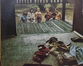 May include: Vintage vinyl record album cover for the Little River Band. The cover features a group of men standing on a wooden structure. Clothing and a hat are scattered on the ground in front of the structure. The album title is displayed at the top.