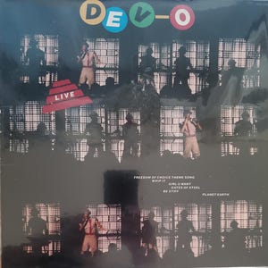 May include: A black and white photo of a band performing live on stage. The band is called DEVO and the photo is from their live album. The album cover features the band's name in large, colorful letters and the word "LIVE" in a red triangle.