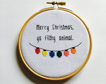 May include: A cross-stitch embroidery hoop with the text "Merry Christmas, ya filthy animal." A string of colorful Christmas lights is stitched below the text. The embroidery is set in a wooden hoop.
