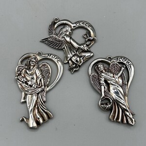 May include: Three silver-toned angel pendants. Each pendant features an angel figure within a heart-shaped frame. The pendants are inscribed with the words "HOPE", "PEACE", and "LOVE".