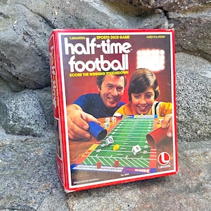 May include: A red cardboard box for Lakeside's Half-Time Football sports dice game. The box cover features an illustration of two people playing the game on a green football field board with white dice, a blue cup, and a red cup.