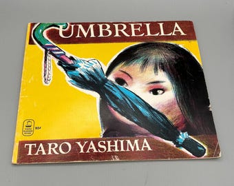 Puede incluir: Una portada de libro infantil vintage con fondo amarillo y el título "Umbrella" en letras rojas grandes y en negrita. La portada presenta una ilustración de una niña sosteniendo una sombrilla azul sobre su rostro. El nombre del autor, "Taro Yashima", está impreso en letras marrones debajo de la ilustración.