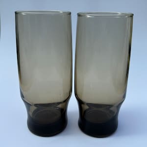 May include: Two tall, vintage, smoky-gray drinking glasses. The glasses have a flared base and a straight, cylindrical body. The base is a darker shade of gray than the body. The glasses are empty and appear to be made of glass.