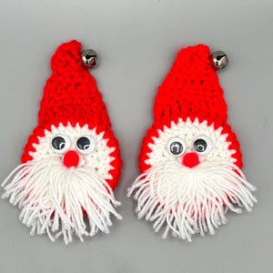 May include: Two handmade crochet ornaments shaped like Santa Claus faces. Each features a bright red pointed hat topped with a small silver metal bell, a white crocheted face, two plastic googly eyes, a red pom-pom nose, and a white yarn beard.